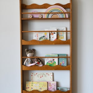 May include: A wooden bookshelf with a wavy design, filled with children's books, toys, and a pastel rainbow. The shelves hold books with titles like "Dear!" and "Princess Poppy." A pink toy car and stuffed animals are also visible.
