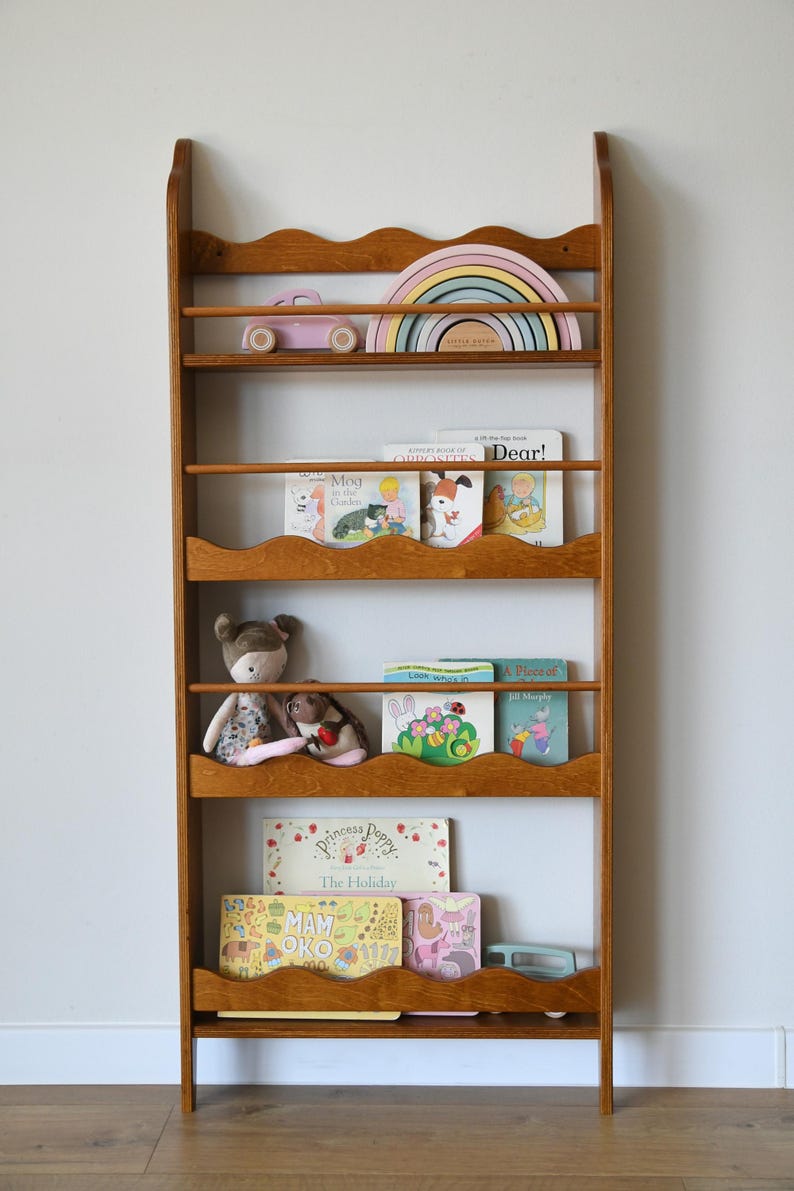 May include: A wooden bookshelf with a wavy design, filled with children's books, toys, and a pastel rainbow. The shelves hold books with titles like "Dear!" and "Princess Poppy." A pink toy car and stuffed animals are also visible.