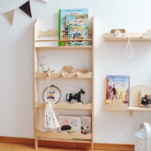 May include: A wooden bookshelf with five shelves, featuring a wavy design on the front edge of each shelf. The shelves are filled with children's books and toys, including a stuffed animal, a wooden rocking horse, and a small bag. The bookshelf is mounted on a white wall with a string of triangle flags hanging above it.