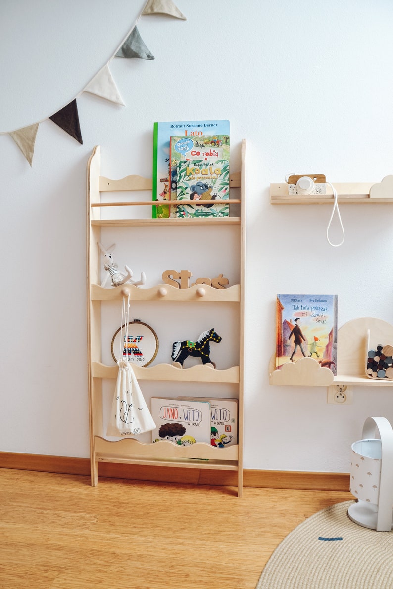 May include: A wooden bookshelf with five shelves, featuring a wavy design on the front edge of each shelf. The shelves are filled with children's books and toys, including a stuffed animal, a wooden rocking horse, and a small bag. The bookshelf is mounted on a white wall with a string of triangle flags hanging above it.