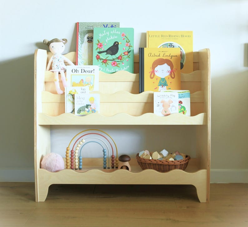 May include: A light wood bookshelf with five shelves, featuring a wavy edge design. The shelves are filled with children's books, a doll, a rainbow abacus, and a basket of colorful wooden blocks.