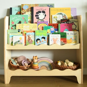 May include: A wooden bookshelf with two shelves, featuring a variety of children's books, including titles like "Coco Chanel", "Little Red Riding Hood", and "Oh Dear!". The bottom shelf has a wooden rainbow toy, a wooden tiger toy, and a wicker basket filled with wooden blocks.
