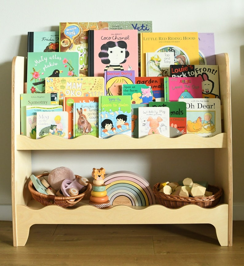 May include: A wooden bookshelf with two shelves, featuring a variety of children's books, including titles like "Coco Chanel", "Little Red Riding Hood", and "Oh Dear!". The bottom shelf has a wooden rainbow toy, a wooden tiger toy, and a wicker basket filled with wooden blocks.