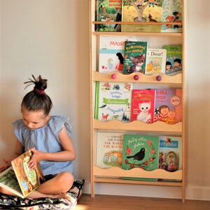 May include: A wooden bookshelf with five shelves, filled with colorful children's books. The bookshelf has a wavy edge and pink knobs. A young girl is sitting on a floral cushion, reading a book.