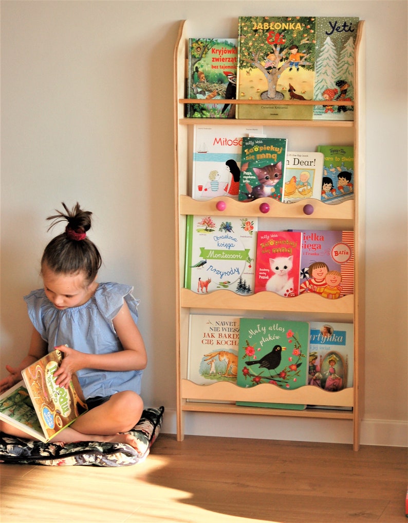 May include: A wooden bookshelf with five shelves, filled with colorful children's books. The bookshelf has a wavy edge and pink knobs. A young girl is sitting on a floral cushion, reading a book.
