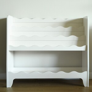 May include: White wooden bookshelf with wavy shelves. The bookshelf has two shelves and is designed to hold books or other items.