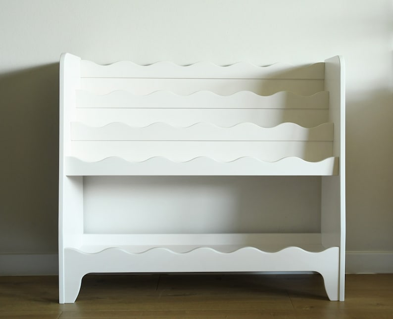 May include: White wooden bookshelf with wavy shelves. The bookshelf has two shelves and is designed to hold books or other items.