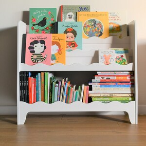 May include: White bookshelf with two shelves, filled with colorful children's books. The books have titles like "Coco Chanel", "Frida Kahlo", "Yeti", "Little Red Riding Hood", and "Wibbly Pig".