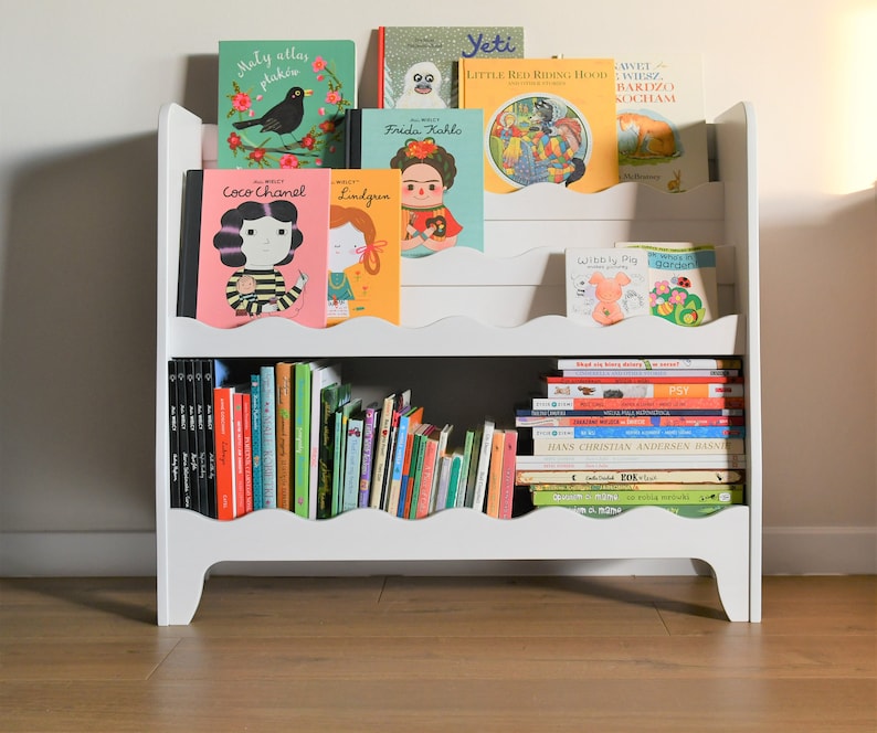 May include: White bookshelf with two shelves, filled with colorful children's books. The books have titles like "Coco Chanel", "Frida Kahlo", "Yeti", "Little Red Riding Hood", and "Wibbly Pig".