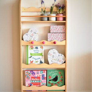 May include: A wooden bookshelf with five shelves, featuring a variety of items, including books, heart-shaped boxes, and potted plants. The bookshelf is painted in a light wood color and has a scalloped edge on the top shelf. The items on the shelves are decorated with floral patterns and pink accents.