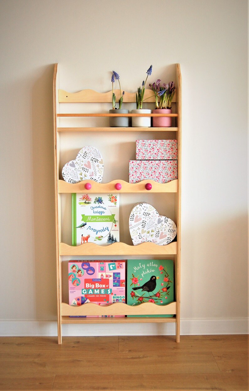 May include: A wooden bookshelf with five shelves, featuring a variety of items, including books, heart-shaped boxes, and potted plants. The bookshelf is painted in a light wood color and has a scalloped edge on the top shelf. The items on the shelves are decorated with floral patterns and pink accents.