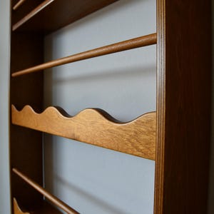 May include: Wooden wall-mounted wine rack with a warm brown finish. The rack features multiple shelves, including a wavy-edged shelf for holding wine bottles and several dowels for hanging glasses. The design is simple and functional.