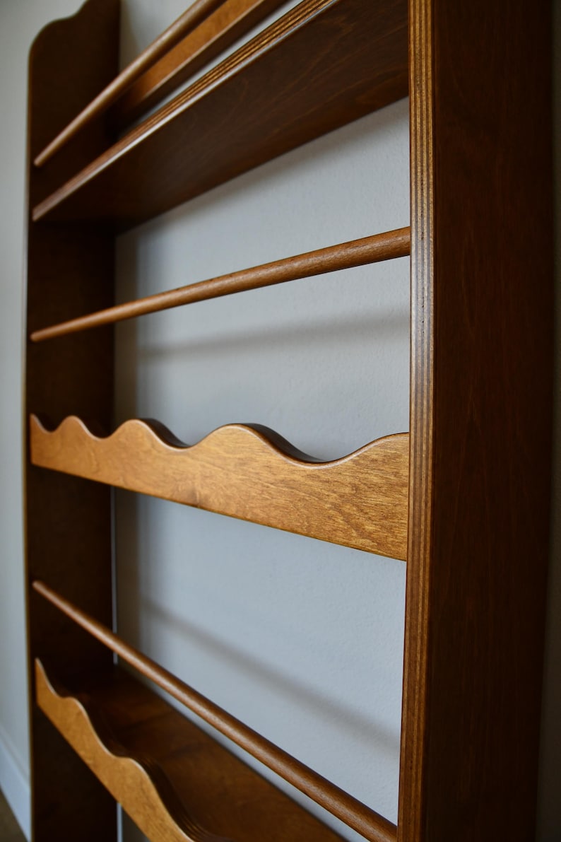 May include: Wooden wall-mounted wine rack with a warm brown finish. The rack features multiple shelves, including a wavy-edged shelf for holding wine bottles and several dowels for hanging glasses. The design is simple and functional.