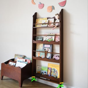 May include: A dark brown wooden bookshelf with multiple shelves, holding children's books and small toys. A smaller, matching wooden book bin is to the left. A decorative fruit garland hangs above.