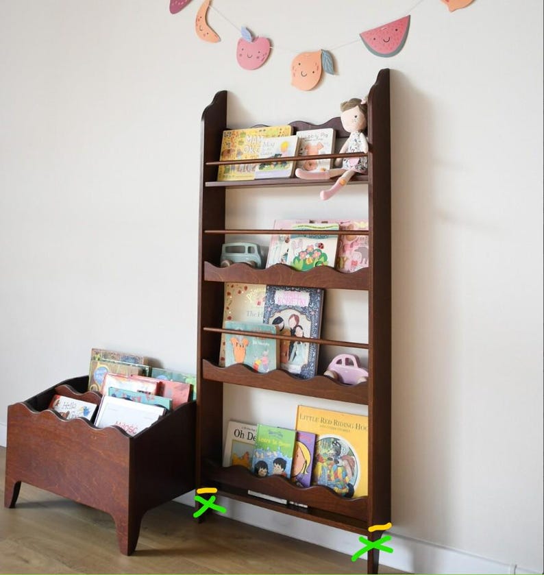 May include: A dark brown wooden bookshelf with multiple shelves, holding children's books and small toys. A smaller, matching wooden book bin is to the left. A decorative fruit garland hangs above.