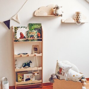 May include: A wooden bookshelf with four shelves, filled with children's books, a stuffed animal, and a toy camera. The bookshelf is against a white wall with two wooden cloud shelves, one with a stuffed sheep and the other with a polka dot container.