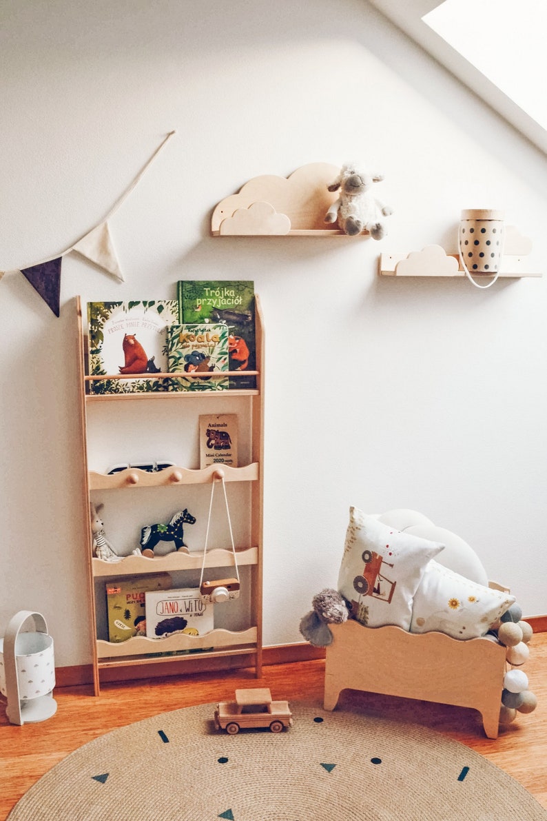 May include: A wooden bookshelf with four shelves, filled with children's books, a stuffed animal, and a toy camera. The bookshelf is against a white wall with two wooden cloud shelves, one with a stuffed sheep and the other with a polka dot container.