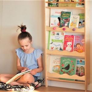 May include: A young girl sits on a floral cushion reading a book. Behind her is a wooden bookshelf with colorful children's books. The books have titles in Polish, including "Mitos", "La&ouml;piekuj sie ming", "ielka Loopiekuj lega", "NAWET NIE WIES JAK BARE CIĘ KOCH", "Maly allas plak&oacute;w", and "Bari tyhka".