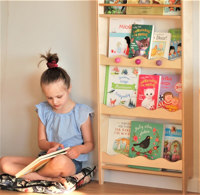 May include: A young girl sits on a floral cushion reading a book. Behind her is a wooden bookshelf with colorful children's books. The books have titles in Polish, including "Mitos", "La&ouml;piekuj sie ming", "ielka Loopiekuj lega", "NAWET NIE WIES JAK BARE CIĘ KOCH", "Maly allas plak&oacute;w", and "Bari tyhka".