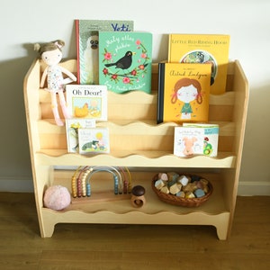 May include: A wooden bookshelf with rounded edges and shelves, filled with children's books. The books have colorful covers with illustrations of animals, people, and objects. There is a doll, a toy abacus, and a basket of colorful blocks on the shelves.
