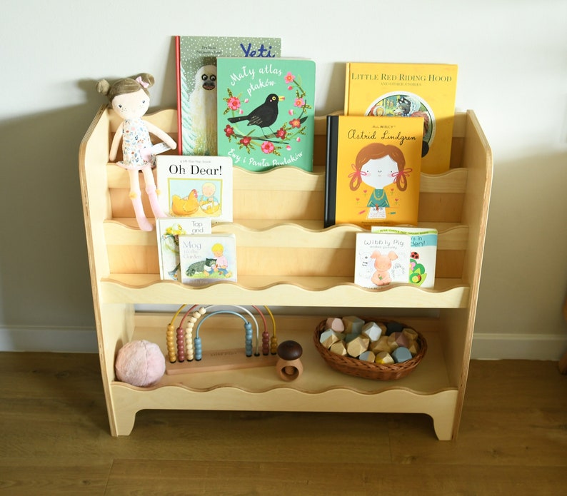May include: A wooden bookshelf with rounded edges and shelves, filled with children's books. The books have colorful covers with illustrations of animals, people, and objects. There is a doll, a toy abacus, and a basket of colorful blocks on the shelves.