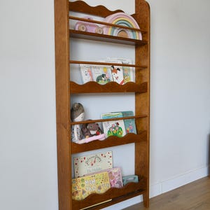 May include: A wooden bookshelf with a warm brown finish. The bookshelf has a scalloped top and several shelves holding children's books, toys, and a wooden rainbow. The bookshelf is against a white wall and a wooden floor.