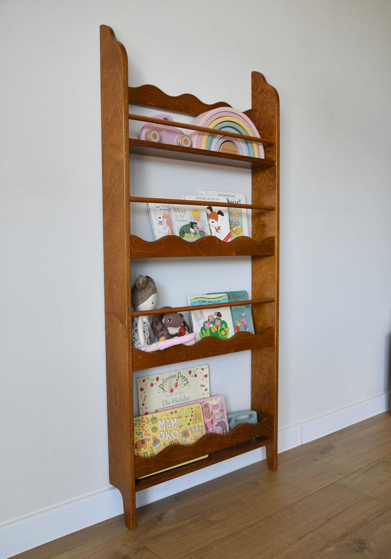 May include: A wooden bookshelf with a warm brown finish. The bookshelf has a scalloped top and several shelves holding children's books, toys, and a wooden rainbow. The bookshelf is against a white wall and a wooden floor.