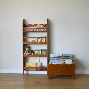 May include: A wooden bookshelf and a matching book bin. The bookshelf has multiple shelves with a scalloped edge design. The shelves hold books, toys, and a rainbow-shaped wooden decoration. The book bin is filled with books.