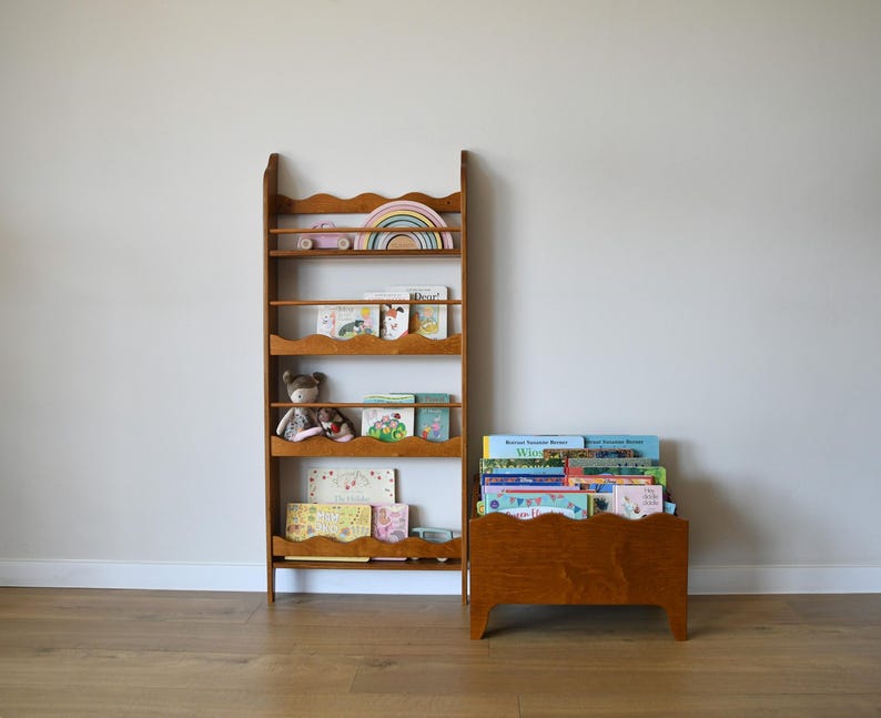 May include: A wooden bookshelf and a matching book bin. The bookshelf has multiple shelves with a scalloped edge design. The shelves hold books, toys, and a rainbow-shaped wooden decoration. The book bin is filled with books.