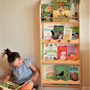 May include: A wooden bookshelf with five shelves, each filled with colorful children's books. The bookshelf is standing in a room with a wooden floor. A young girl is sitting on a floral cushion, reading a book.