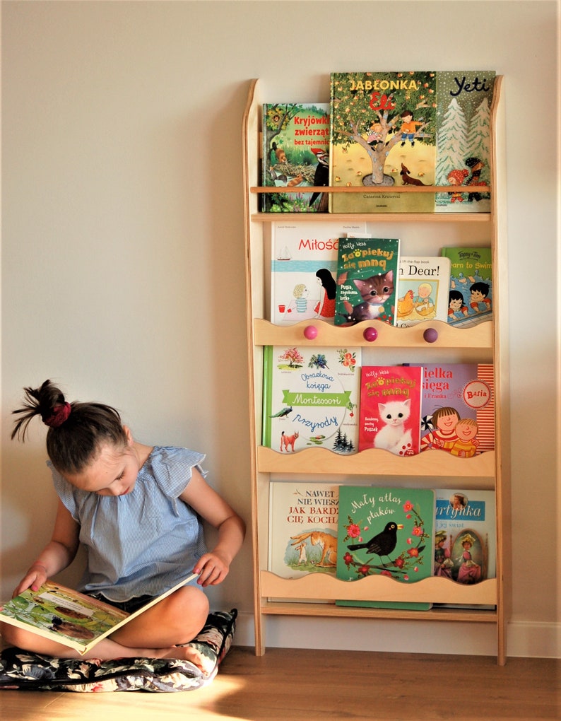May include: A wooden bookshelf with five shelves, each filled with colorful children's books. The bookshelf is standing in a room with a wooden floor. A young girl is sitting on a floral cushion, reading a book.