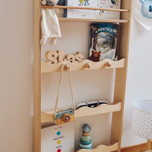 May include: A wooden bookshelf with four shelves, featuring a variety of children's books, a stuffed animal, a toy camera, and a wooden stacking toy. The bookshelf is painted in a light wood color and has a wavy design on the edges of the shelves.