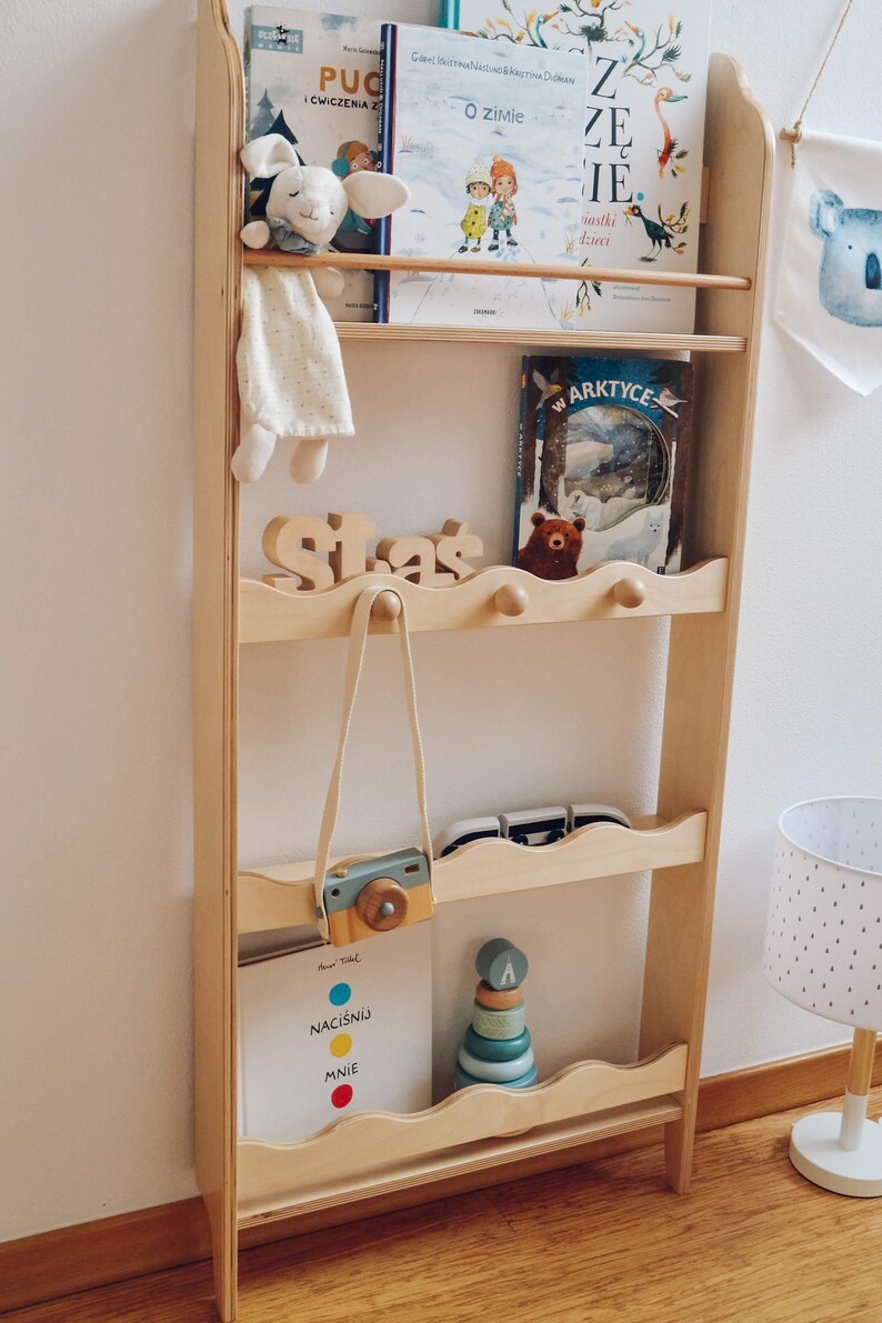 May include: A wooden bookshelf with four shelves, featuring a variety of children's books, a stuffed animal, a toy camera, and a wooden stacking toy. The bookshelf is painted in a light wood color and has a wavy design on the edges of the shelves.