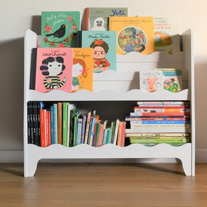 May include: White bookshelf with two shelves, filled with colorful children's books. The books have titles like "Coco Chanel", "Frida Kahlo", "Yeti", "Little Red Riding Hood", and "Wibbly Pig".