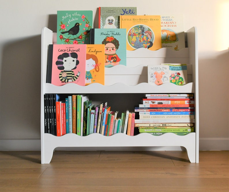 May include: White bookshelf with two shelves, filled with colorful children's books. The books have titles like "Coco Chanel", "Frida Kahlo", "Yeti", "Little Red Riding Hood", and "Wibbly Pig".