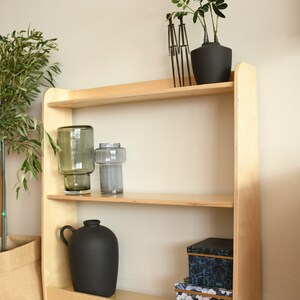 May include: A light wood bookshelf with three shelves. The top shelf has a potted plant with green leaves and a black pot. The middle shelf has two clear glass vases and the bottom shelf has a black vase and two boxes with floral patterns.