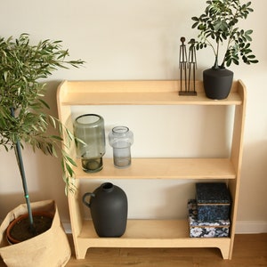 May include: A light wood shelf unit with three shelves. The top shelf has a small metal sculpture and a black vase with a green plant. The middle shelf has two glass vases. The bottom shelf has a black vase and two boxes with floral patterns.