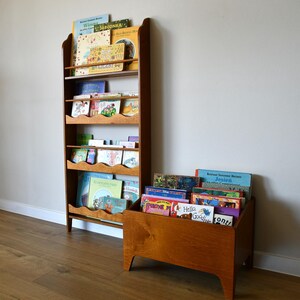 May include: Wooden bookshelf and a matching storage box filled with children's books. The bookshelf has multiple shelves with a scalloped edge design. The books are colorful and varied, suggesting a children's reading collection.