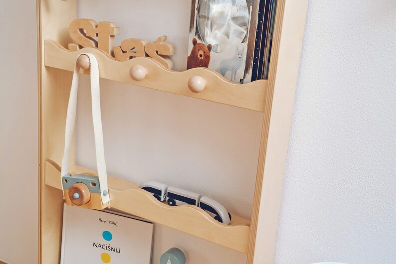 May include: A wooden shelf unit with three shelves, each with a decorative wave-shaped edge. The top shelf has a wooden sign that says "Stars" and a stuffed bear. The middle shelf has a wooden camera with a strap hanging from a hook. The bottom shelf has a book with the title "Naci&scaron;ni&uacute;" and a toy car.