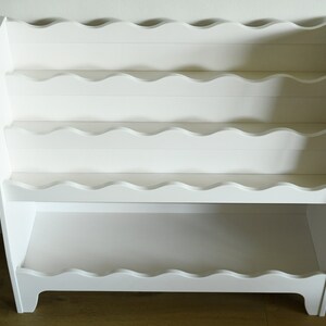 May include: White wooden bookshelf with scalloped edges on the shelves. The bookshelf has four shelves and stands on four legs.