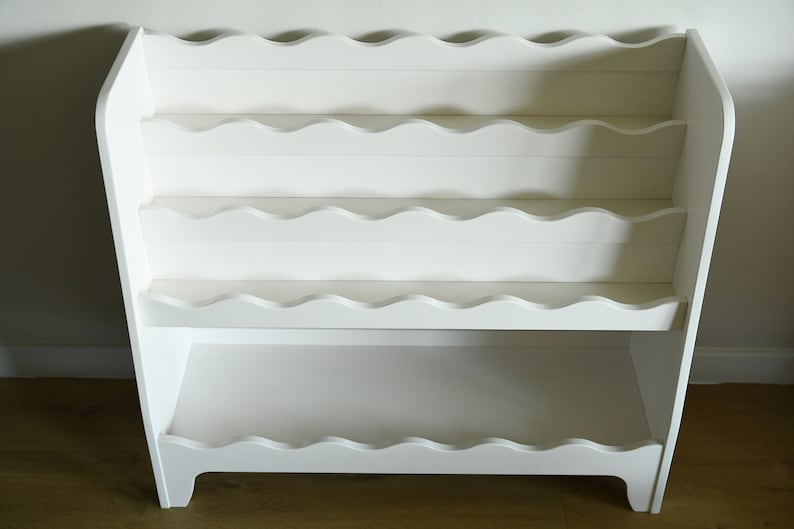 May include: White wooden bookshelf with scalloped edges on the shelves. The bookshelf has four shelves and stands on four legs.