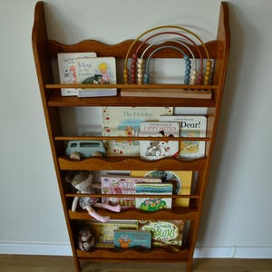May include: A wooden bookshelf with a scalloped top, filled with children's books and toys. The shelf is a warm brown color and has multiple levels. A wooden abacus with colorful beads sits on the top shelf. A stuffed doll and toy car are also visible.