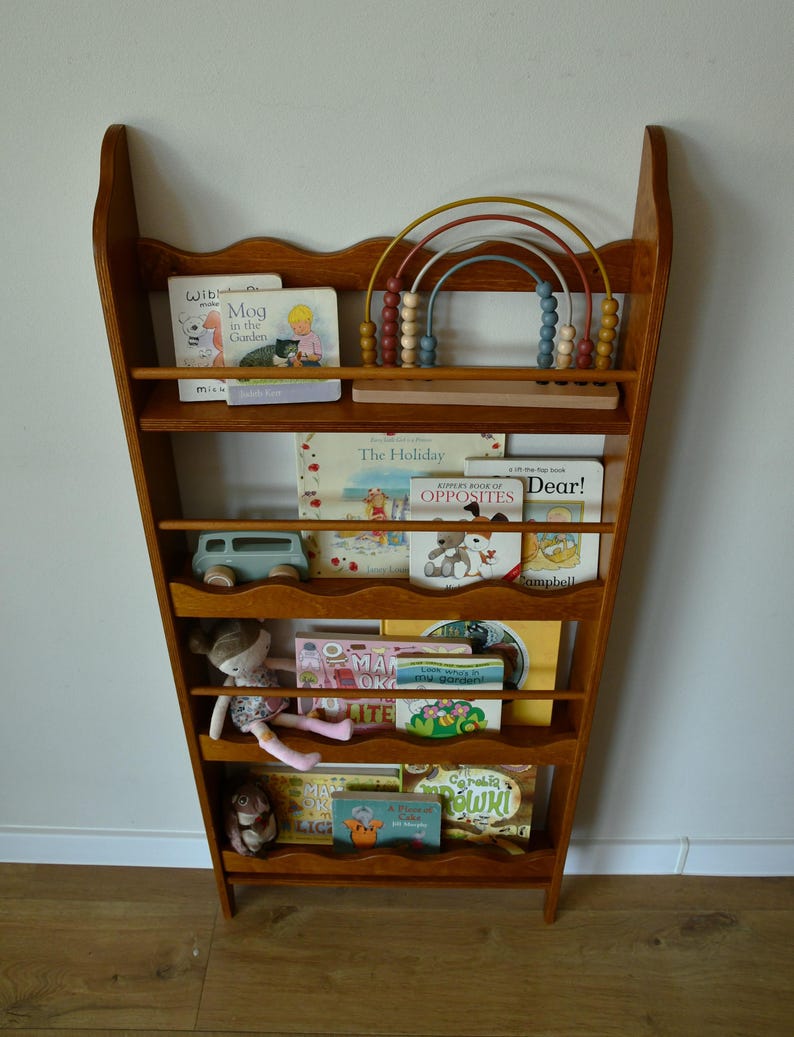 May include: A wooden bookshelf with a scalloped top, filled with children's books and toys. The shelf is a warm brown color and has multiple levels. A wooden abacus with colorful beads sits on the top shelf. A stuffed doll and toy car are also visible.