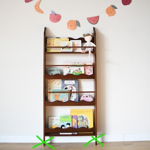 May include: A dark brown wooden bookshelf filled with children's books. A small doll sits on the top shelf. A decorative fruit garland hangs above the bookshelf. Two toy cars are on the shelves. The bookshelf has a scalloped edge.