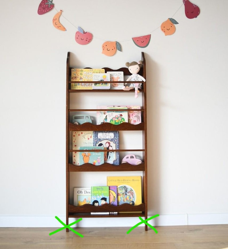 May include: A dark brown wooden bookshelf filled with children's books. A small doll sits on the top shelf. A decorative fruit garland hangs above the bookshelf. Two toy cars are on the shelves. The bookshelf has a scalloped edge.