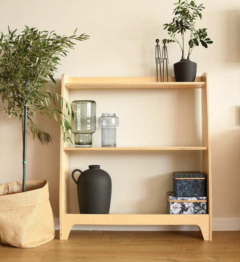 May include: A light wood shelf unit with two shelves. The top shelf has a small metal sculpture, a green glass vase, and a potted plant with green leaves. The bottom shelf has a black ceramic vase, two boxes with floral patterns, and a potted plant with green leaves.