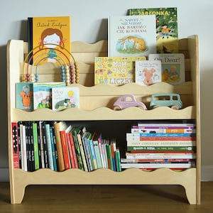May include: A wooden bookshelf with multiple shelves, featuring a variety of children's books, including titles like "Wibbly Pig" and "Oh Dear!" The bookshelf has a wavy design on the edges of the shelves and is filled with books of different sizes and colors.