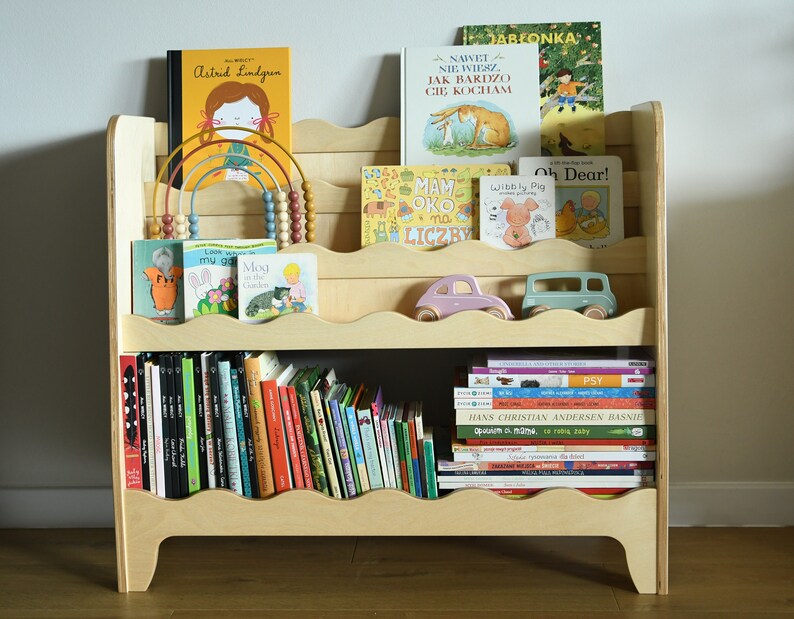 May include: A wooden bookshelf with multiple shelves, featuring a variety of children's books, including titles like "Wibbly Pig" and "Oh Dear!" The bookshelf has a wavy design on the edges of the shelves and is filled with books of different sizes and colors.