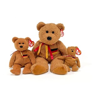 May include: Three brown teddy bears of varying sizes. Each bear has a black nose, a red and white tag, and a small German flag patch. The largest bear has a ribbon with the German flag colors.
