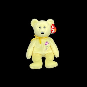 May include: A soft, yellow teddy bear with a purple flower embroidered on its chest. The bear has a yellow ribbon around its neck and a red tag attached to its ear. The bear is a collectible item.